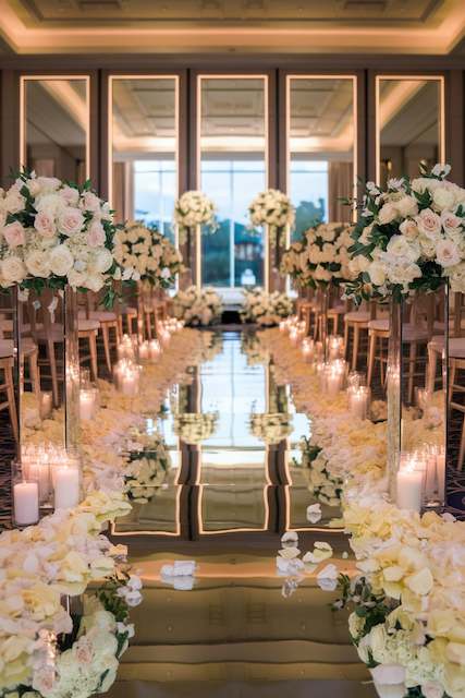 Golden Mirror Aisle A lavish wedding aisle features a gold reflective floor surrounded by white rose petals and candles. Tall floral arrangements with white roses line the aisle, creating an elegant and glamorous atmosphere in a grand venue.
