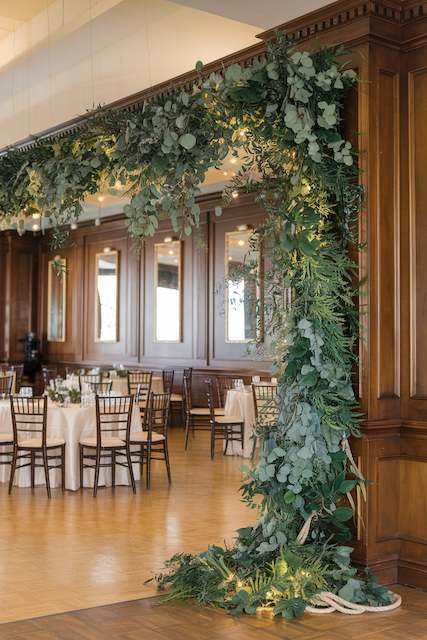 Greenery Garlands An archway of lush greenery adorns a wooden interior, creating an elegant and natural entrance to a dining area with round tables.