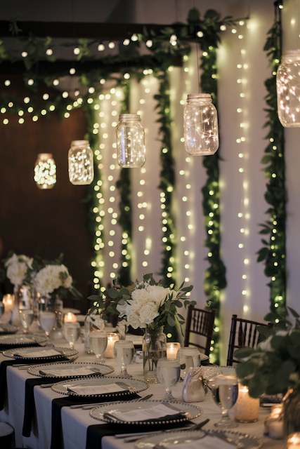 Hanging Mason Jar Lights Mason jars filled with string lights hang above a long table set with white floral centerpieces and elegant tableware, creating a warm and romantic atmosphere.
