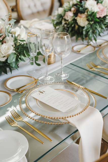 Layered Glass Charger Plates Elegant table setting with glass plates featuring gold rims, a napkin draped over the edge, and surrounded by gold flatware and glassware. Floral arrangements add a lush, romantic touch.