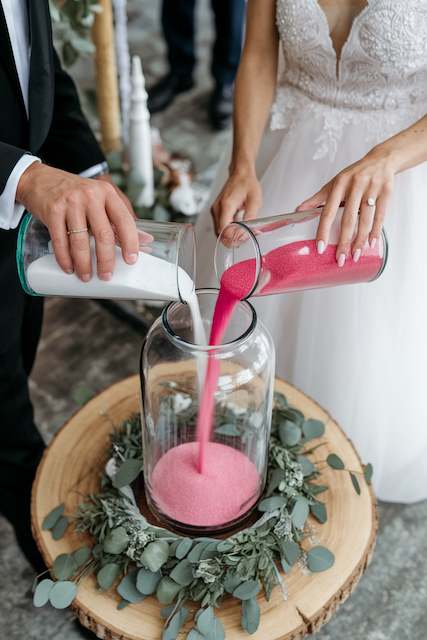 Sand Ceremony Bride and groom pour white and pink sand into a jar during a unity sand ceremony on a wood slice altar edged with eucalyptus.