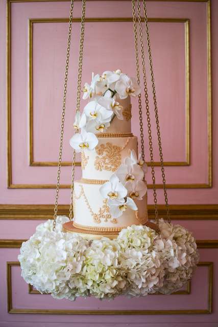 Suspended Cake Installation with Gold Accents A tiered wedding cake with gold detailing and white orchids is suspended by chains, surrounded by white hydrangeas, creating an elegant display against a pink backdrop.