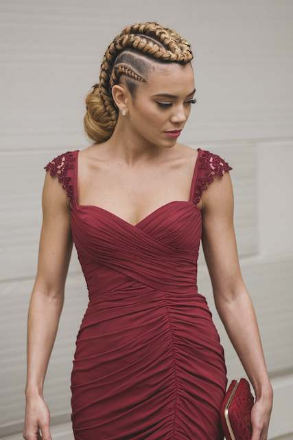 Braided Mohawk Updo – A bold and edgy updo featuring a central braid. Bridesmaid with a bold braided mohawk updo and side cornrows wearing a deep burgundy lace dress, posing outdoors against a neutral wall.