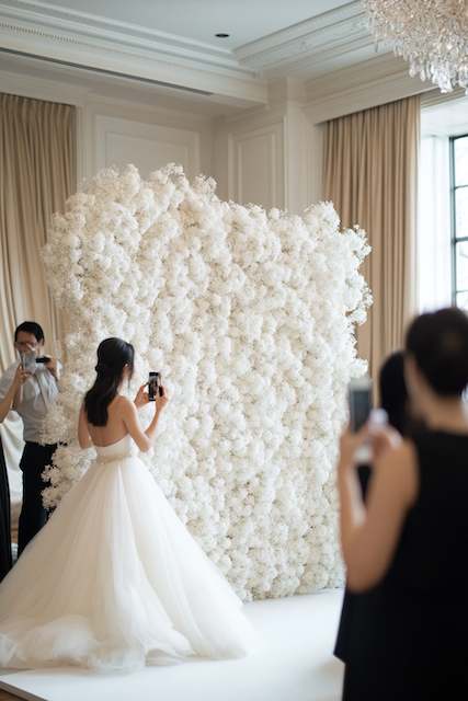 Breathtaking Baby's Breath Backdrop Bride in a tulle gown photographs a towering baby's breath flower wall as guests capture the moment inside a bright ballroom