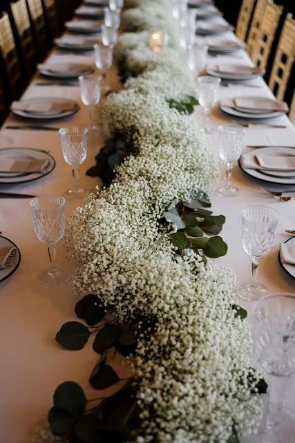 Cascading Baby’s Breath Waterfall Centerpiece A long banquet table adorned with a lush garland of baby’s breath and eucalyptus, creating a soft and elegant centerpiece for a wedding reception