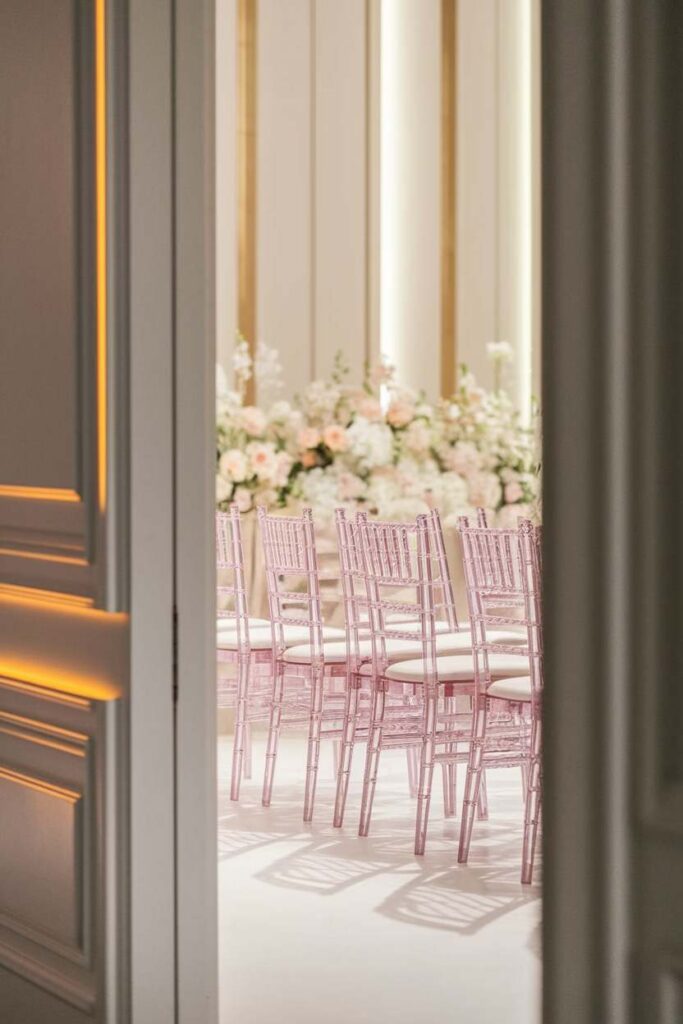 Ceremony Flow and Key Logistics Elegant ceremony setup with soft pink chairs and white florals viewed through open ballroom doors