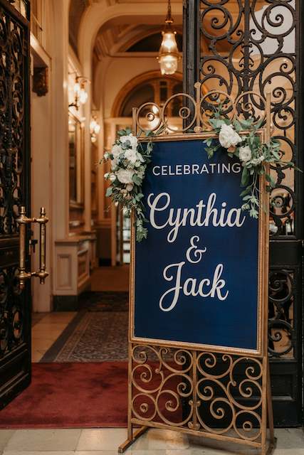 Custom Welcome sign as classy wedding decor Navy wedding welcome sign reading “Celebrating Cynthia and Jack” on an ornate gold stand with white florals at an elegant entry.