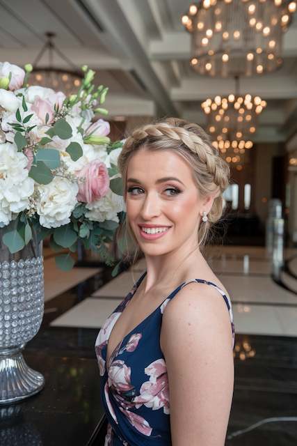 Dutch Braid Crown Bridesmaid with a Dutch braid crown and soft tendrils posing beside a large blush and white floral arrangement in an elegant ballroom.