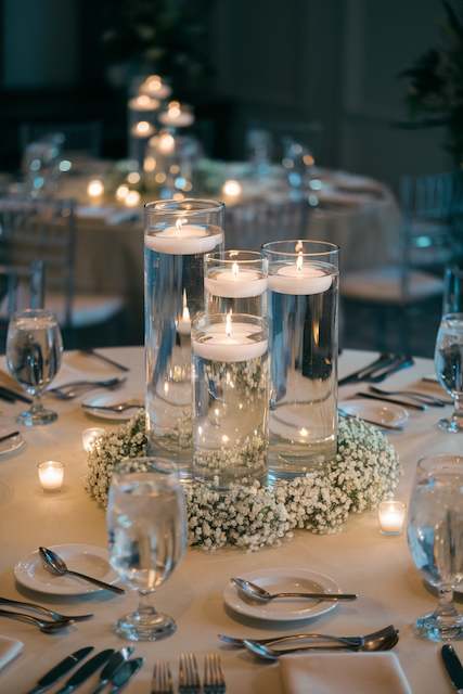 Floating Candles with Baby’s Breath Rings Romantic wedding table centerpiece featuring floating candles arranged in tall glass cylinders, surrounded by delicate baby’s breath, creating a soft and elegant ambiance