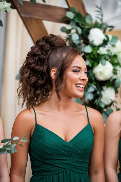 High-Volume Textured Ponytail Bridesmaid with a high volume textured ponytail and soft face framing strands wearing a green dress, smiling at an outdoor wedding ceremony.