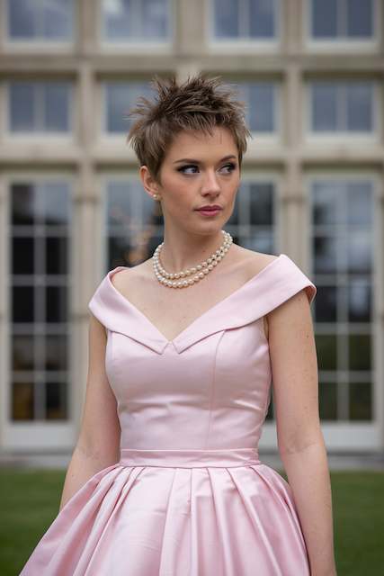 Short and Spiky Bridesmaid with a short spiky pixie cut wearing a pale pink off shoulder dress and pearl necklace, standing outdoors in front of tall windows.