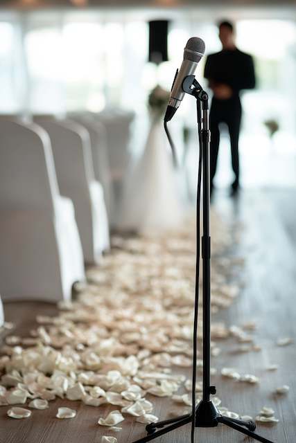 Microphone on stand along ceremony aisle with white chairs and rose petals, officiant blurred in background before vows. Microphone on stand along ceremony aisle with white chairs and rose petals, officiant blurred in background before vows.