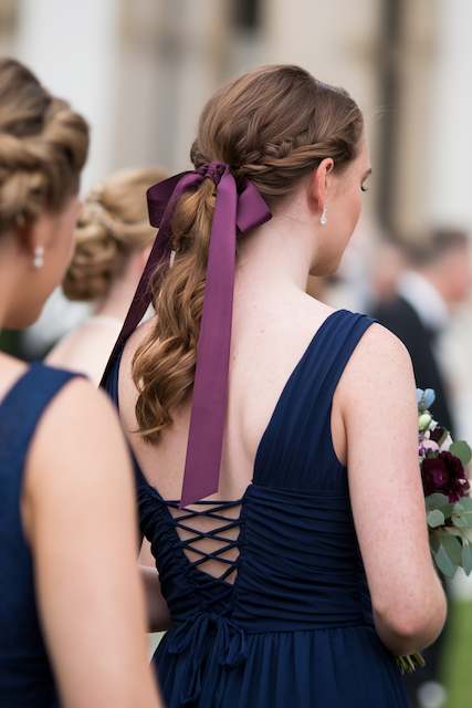 Twisted Ponytail with Ribbon Bridesmaid with a twisted ponytail tied with a long plum satin ribbon, wearing a navy dress and holding flowers at an outdoor wedding.