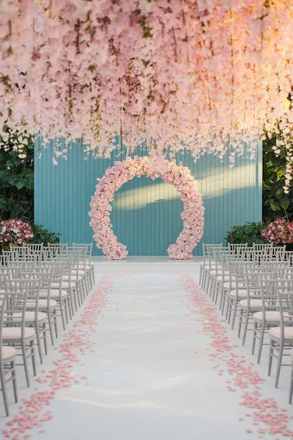 Wedding Ceremony Budget Outdoor ceremony with silver chairs, petal lined aisle, and a round blush floral arch beneath cascading pink blooms and a teal backdrop