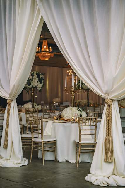 White Linen Drapes With Gold Rope Tiebacks. Elegant reception space with white draped curtains tied with gold rope, revealing tables set with gold Chiavari chairs, white linens, and lush floral centerpieces. Chandeliers add a warm glow.