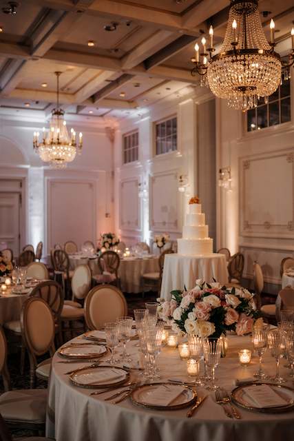 classy wedding decor with Wall uplighting in soft colors Elegant ballroom reception with crystal chandeliers, blush-toned tables, white and blush rose centerpiece, gold-rim chargers, glowing votives, and a tiered wedding cake on display.