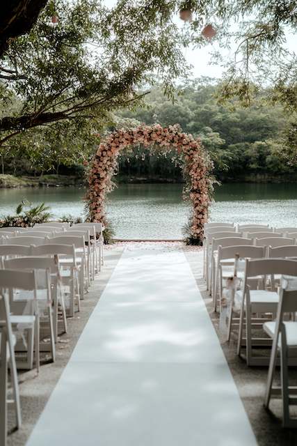 folding chairs for ceremony Lakeside ceremony aisle with white runner and rows of white chairs leading to a blush floral arch beneath shady trees by calm water.