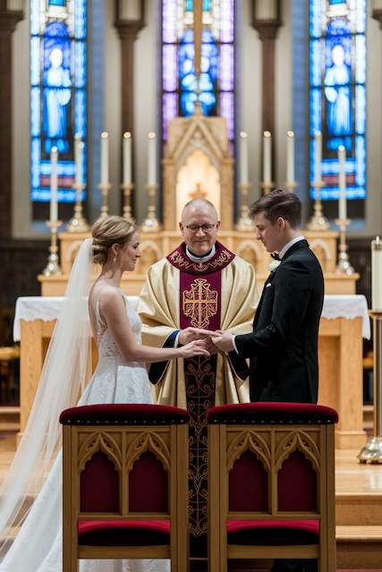 religious wedding ceremony Bride and groom exchange rings before a smiling priest at the altar, framed by stained glass windows and tall candles in a grand church.