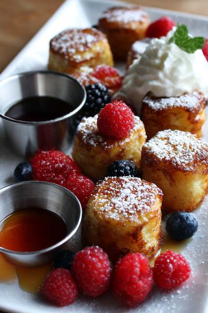 French Toast Bites Mini French toast bites topped with fresh berries powdered sugar and maple syrup served with whipped cream for a brunch wedding dessert station.