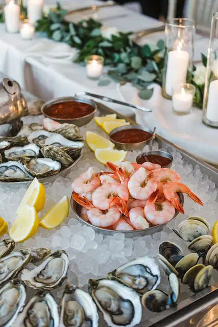 Seafood Sensations Raw bar display with shrimp, oysters, and clams on ice, lemon wedges, and cocktail sauce; candles and greenery behind