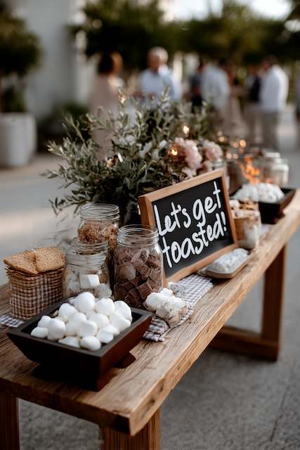 S’mores bar A rustic s'mores bar setup with marshmallows, graham crackers, and chocolate chunks in jars, accompanied by a 'Let's Get Toasted' sign on a wooden table surrounded by greenery and soft lighting