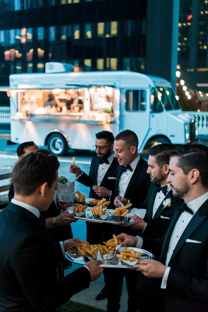 a-luxurious-event-at-a-hotel-with-a-slee_iaV5w_YCR7yjxf34_PWWAQ_xhnQS55uTDK0vg2s8mBZmA Groomsmen in tuxedos share trays of fries by a white food truck at an outdoor wedding reception, city lights behind.
