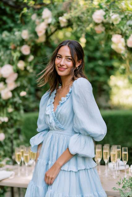 Bridesmaid wearing puffy sleeves dress at rehearsal dinner Bridesmaid wearing a light blue puff-sleeve dress, standing outdoors in front of a floral arch, smiling at the rehearsal dinner event