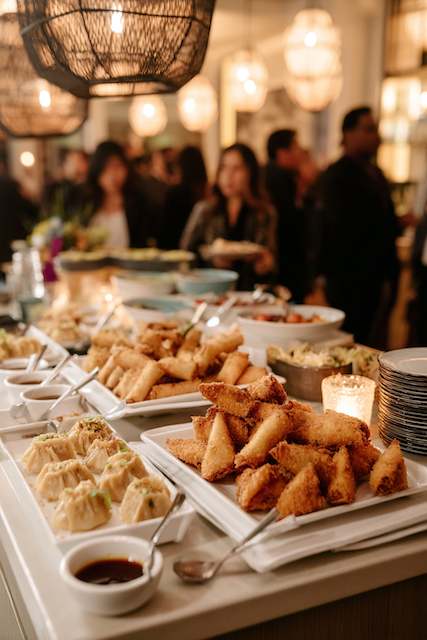 Dumpling and Spring Roll Bar Dumplings and spring rolls elegantly arranged on trays with dipping sauces at a lively wedding reception, creating a vibrant atmosphere.