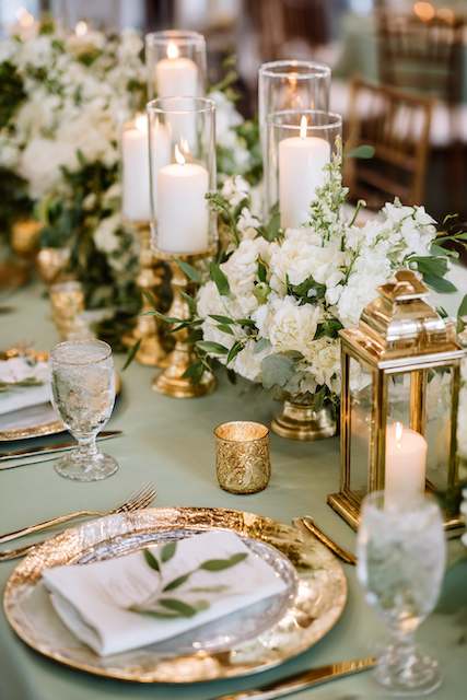 Sage Green & Gold wedding dinner table setting Elegant sage green and gold wedding tablescape featuring gold-rimmed chargers, glassware, ivory floral centerpieces, tall candles in glass holders, and gold lanterns for a warm, romantic glow