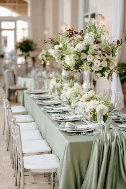 Sage Green & Silver wedding dinner table Elegant wedding table setting with a sage green table runner, white floral centerpiece, and lush greenery, set outdoors with natural light