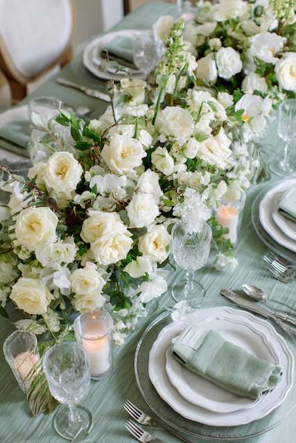 Sage Green & White wedding dinner table setting Elegant wedding reception table with a sage green runner, white rose floral arrangements, candles, and fine china in a soft and romantic setting