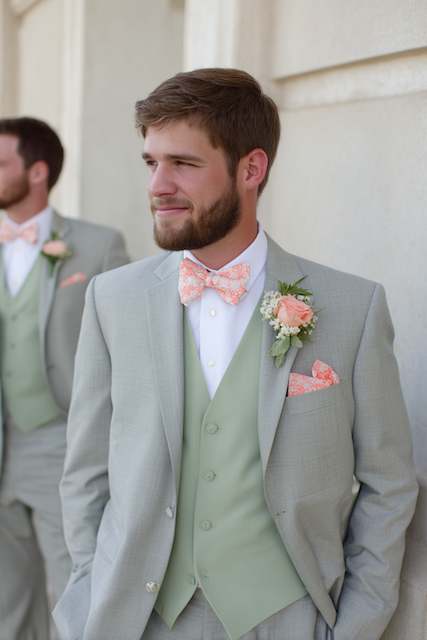 groomsmen in sage green suits Groomsman wearing a sage green suit with a peach bow tie, pocket square, and boutonnière, standing with hands in pockets