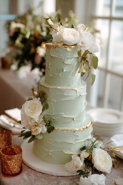 sage green frosted cake with gold accents Three-tier sage green wedding cake with textured buttercream, gold leaf accents, and white floral decorations featuring roses and eucalyptus leaves