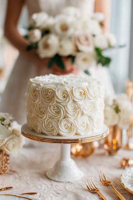 All-white rosette masterpiece wedding cake white wedding cake with intricate buttercream rosette decorations, set on a delicate cake stand, surrounded by soft floral arrangements and golden accents