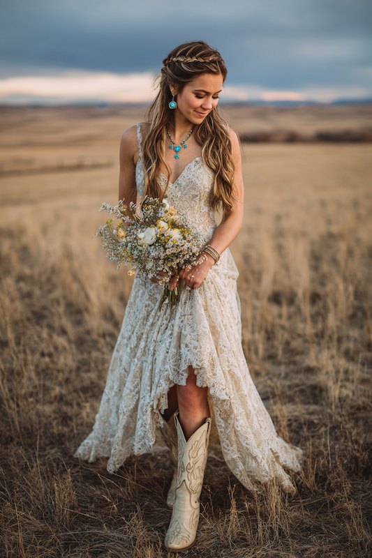 A bride in a lace wedding dress with cowboy boots, holding a bouquet, smiling in a rustic outdoor setting