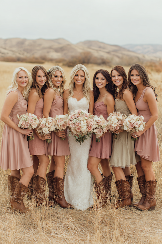 A bride and her bridesmaids, all in cowboy boots, standing together holding bouquets in a rustic outdoor setting