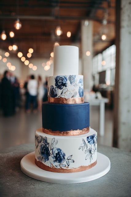 Navy + copper wedding cake Four tier wedding cake with alternating tiers of white and deep navy fondant, featuring hand-painted blue floral patterns and metallic copper bands, blending modern elegance with artistic flair.