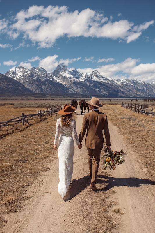 A bride and groom walk hand-in-hand on a dirt path with mountains and horses in the background.