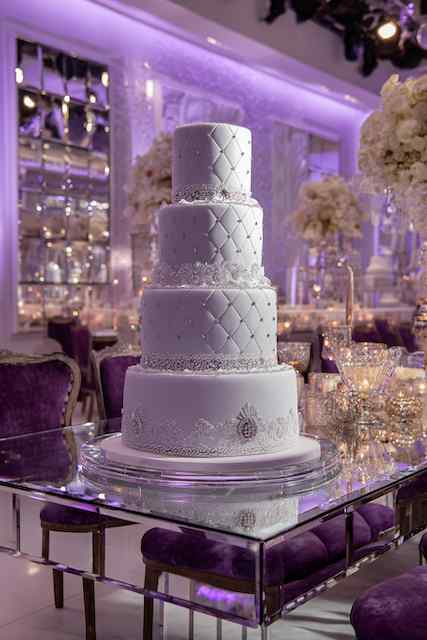 Quilted fondant with brooch detail wedding cake Four-tiered wedding cake with quilted fondant, pearl detailing, and intricate lace trim, set on a clear pedestal with a purple and elegant reception backdrop