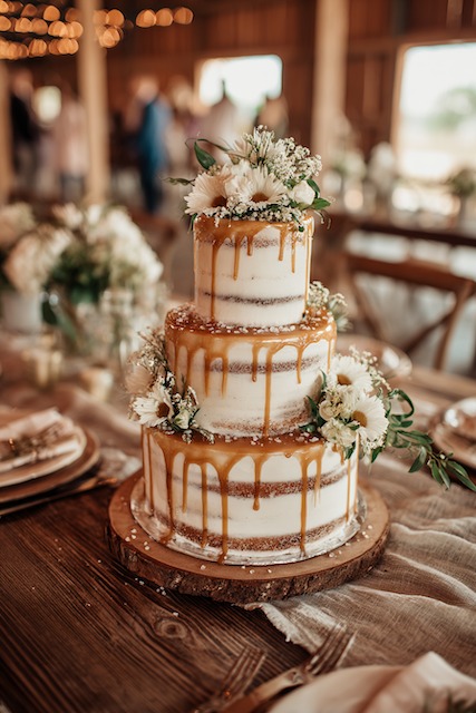 Salted caramel + vanilla wedding cake Three tier semi-naked wedding cake drizzled with golden caramel, topped and adorned with white daisies, baby's breath, and greenery, creating a cozy rustic charm perfect for barn or outdoor celebrations.