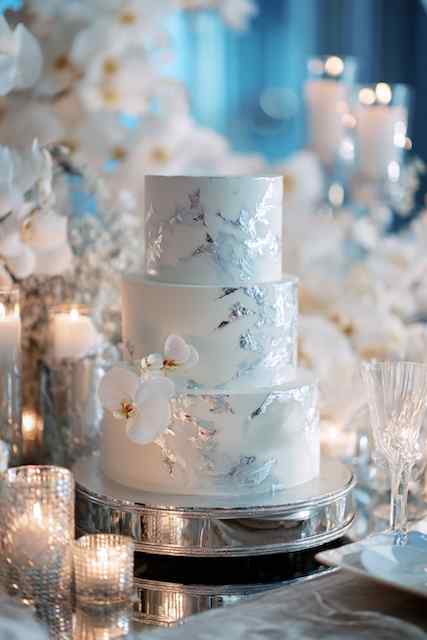 Silver-stacked tiers wedding cake three-tiered wedding cake with a sleek marble effect, decorated with delicate white orchids and set against a soft, blurred background of candles and floral arrangements