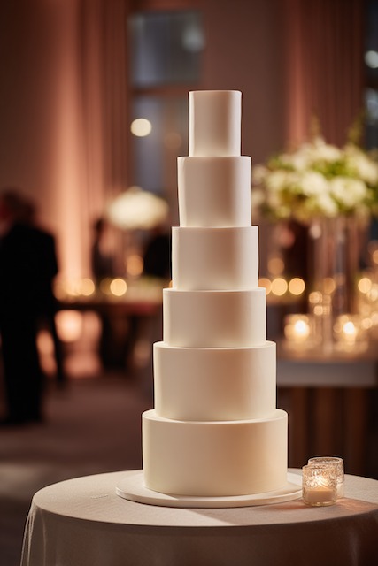 Tall skinny tiers wedding cake Tall seven-tier white wedding cake with smooth fondant finish, minimalist design, and clean modern lines, displayed in a softly lit elegant reception setting.