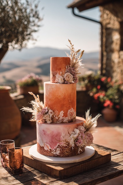 Terracotta + rust wedding cake Three tier terracotta ombré wedding cake with warm rust and blush tones, adorned with dried florals, pampas grass, and roses, exuding rustic Tuscan elegance in golden outdoor light.