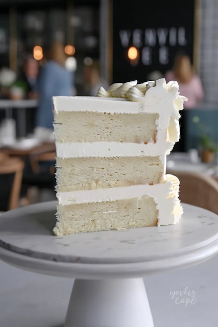 Vanilla bean almond wedding cake Three layer vanilla bean wedding cake slice with silky white buttercream frosting and light, fluffy sponge, garnished with delicate white chocolate shavings on top for an elegant minimalist presentation.