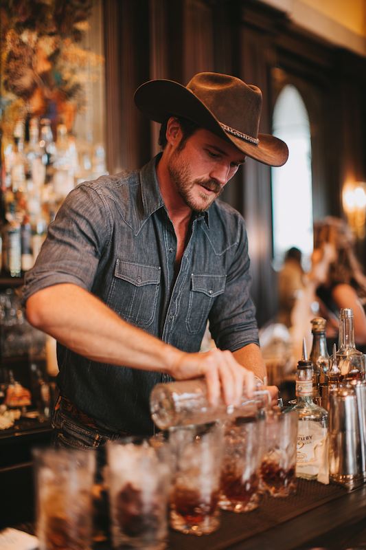 A bartender in cowboy attire mixing drinks at a cowboy-themed wedding reception, with a stylish bar setup and guests in the background