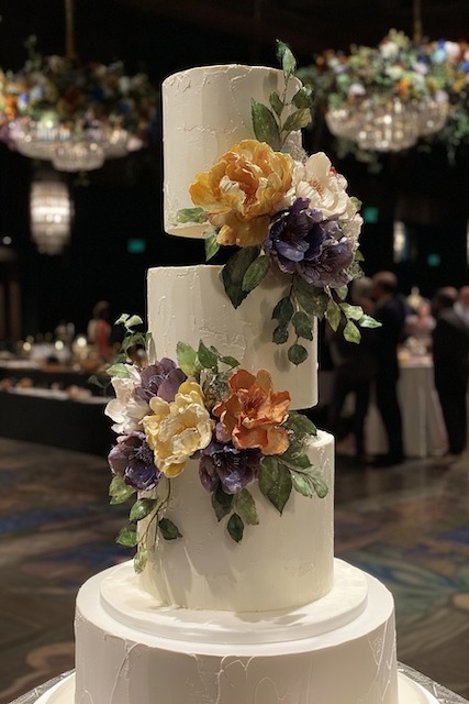 floating tiers wedding cake Three tier round wedding cake with smooth white buttercream and bold handcrafted sugar flowers in deep purple, gold, and ivory, creating a modern elegant contrast with lush green leaves.