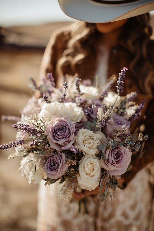 A bride in a western-style cowboy hat holding a beautiful bouquet of lavender, pale roses, and soft greenery, capturing the essence of rustic elegance for her cowboy wedding