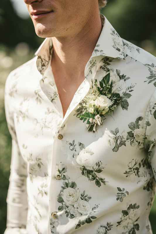 groom ditching the tie floral button-down shirt Close-up of a groom wearing a floral button-down shirt with a boutonnière featuring white roses and greenery, perfect for a casual, country-themed wedding