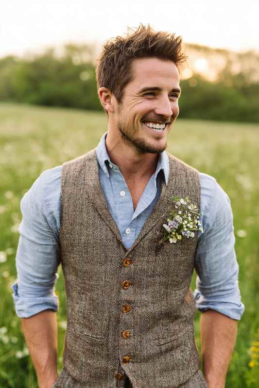 groom in Chambray shirt with tweed vest Groom wearing a chambray shirt with a tweed vest, smiling with a boutonnière, set against a natural outdoor backdrop