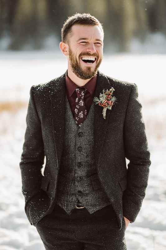 groom in Dark suits in warm fabrics A groom smiling in the snow, dressed in a dark brown tweed suit, burgundy shirt, floral tie, and rustic boutonnière, perfect for a winter country-themed wedding